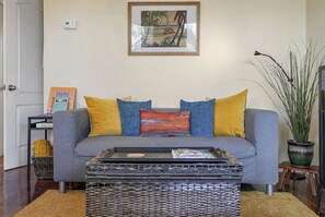 Una sala de estar con un sofá gris, cojines amarillos y azules, una mesa de centro de mimbre, un cuadro en la pared y una planta en maceta.