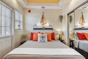 A hotel room with a bed, a nightstand, a lamp, a wall-mounted shelf with plants, and a window with blinds.
