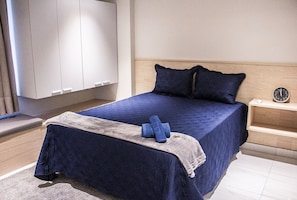 Un lit bien fait, avec une couvre-lit bleue et des oreillers, une table de chevet en bois avec une horloge, et une grande armoire.