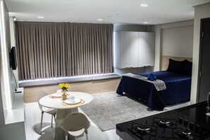 A modern hotel room with a bed, a dining area with a table and chairs, a kitchen area with a stove, and a large window with curtains. - Apart no Maradisa Design por Carpediem (João Pessoa)