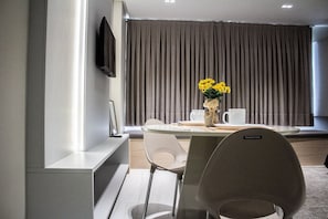 Une chambre d’hôtel moderne dotée d’un coin repas comprenant une table ronde, deux chaises, un vase avec des fleurs jaunes et un téléviseur à écran plat fixé au mur.