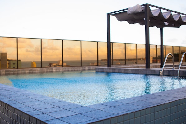 Une piscine sur le toit, entourée d’une paroi en verre, avec un espace salon couvert et une vue sur le paysage urbain.