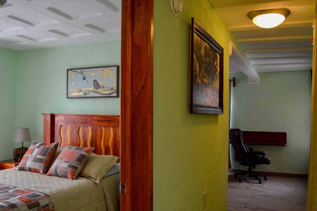 Une chambre à coucher avec une tête de lit en bois, un lit recouvert d’une literie à motifs, une table de chevet avec une lampe et un tableau encadré accroché au mur.