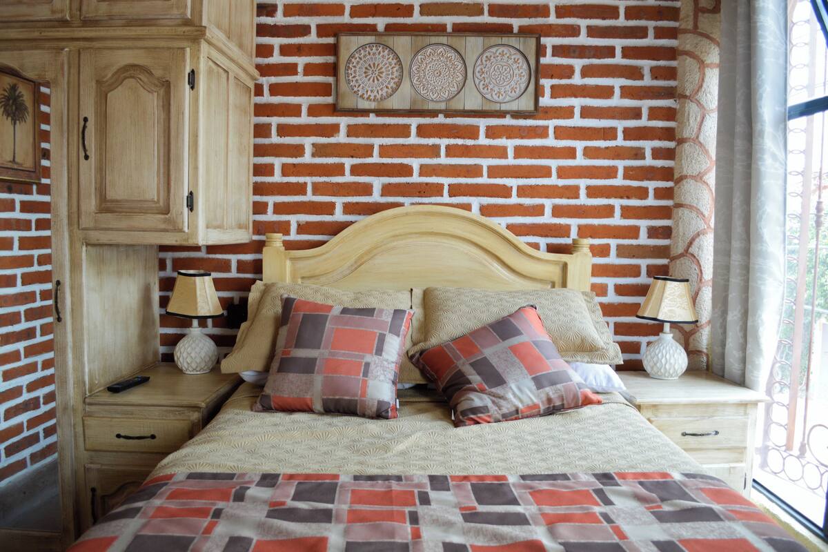 Une chambre avec un mur en briques, une tête de lit en bois, deux tables de chevet avec des lampes, un lit avec une literie à motifs et un élément décoratif mural au-dessus du lit.