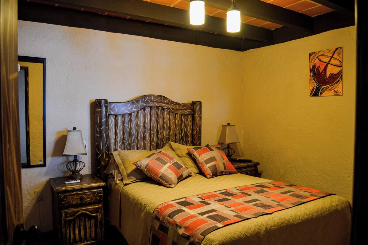 Une chambre à coucher avec une tête de lit en bois, deux lampes de chevet, une couvre-lit matelassé et une œuvre d’art encadrée accrochée au mur.
