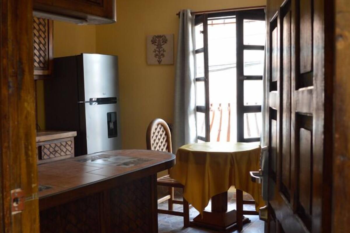 Une petite cuisine comprenant un réfrigérateur, une table à manger avec des chaises et une fenêtre avec des rideaux.