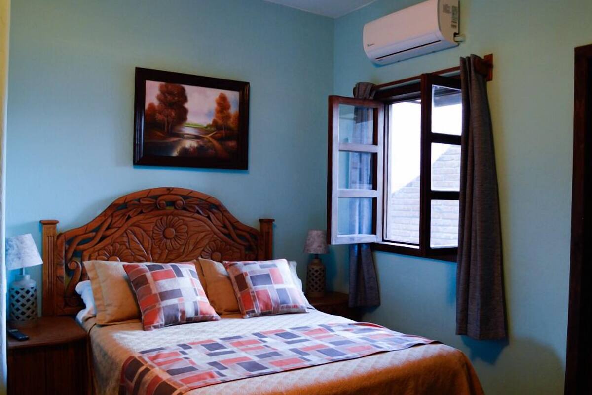Une chambre à coucher avec une tête de lit en bois, un lit recouvert d’une couvre-lit à motifs, un tableau accroché au mur et une fenêtre ouverte avec des rideaux.