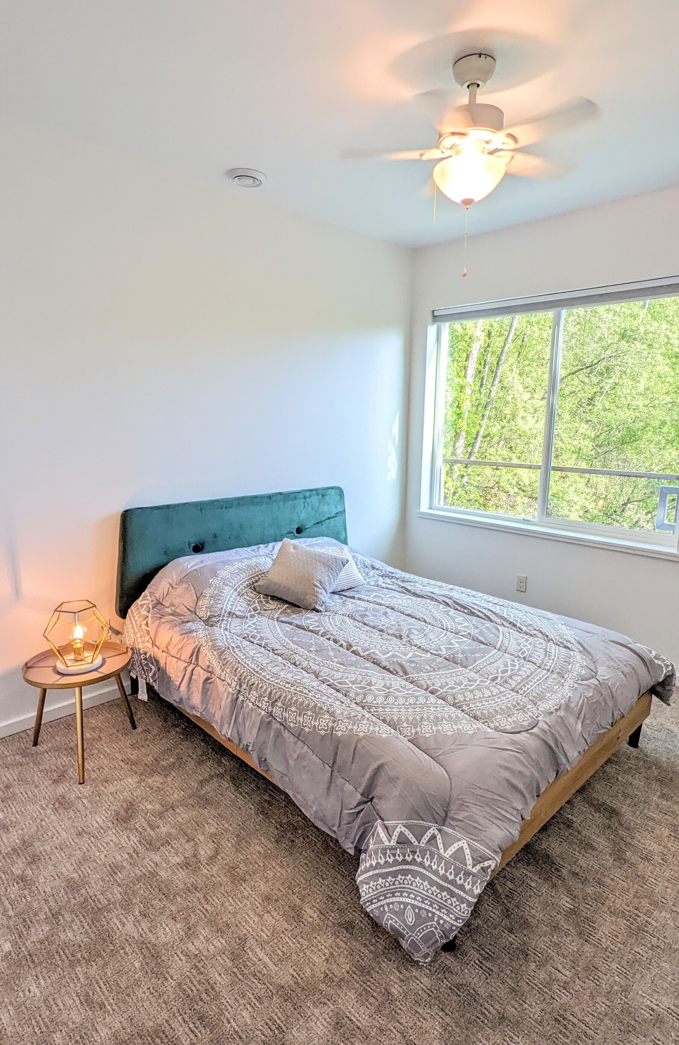 A bedroom with a bed, a ceiling fan, a window, and a nightstand with a lamp.