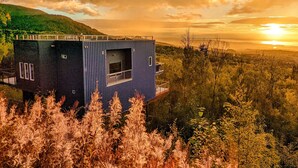 A modern, multi-level cabin with a balcony, surrounded by forested hills, during sunset. - Beautiful Modern Loft | Denali Views, Pool Table, Roof Deck, Firepit (Anchorage)