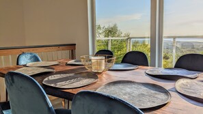 Een eethoek met een houten tafel en stoelen, een geweven decoratiestuk in het midden, en een uitzicht op groen en verre bergen door grote ramen.