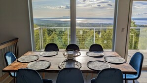 A dining table with six chairs and a central candle holder, set against a large window overlooking a scenic landscape with trees and a body of water. - Beautiful Modern Loft | Denali Views, Pool Table, Roof Deck, Firepit (Anchorage)