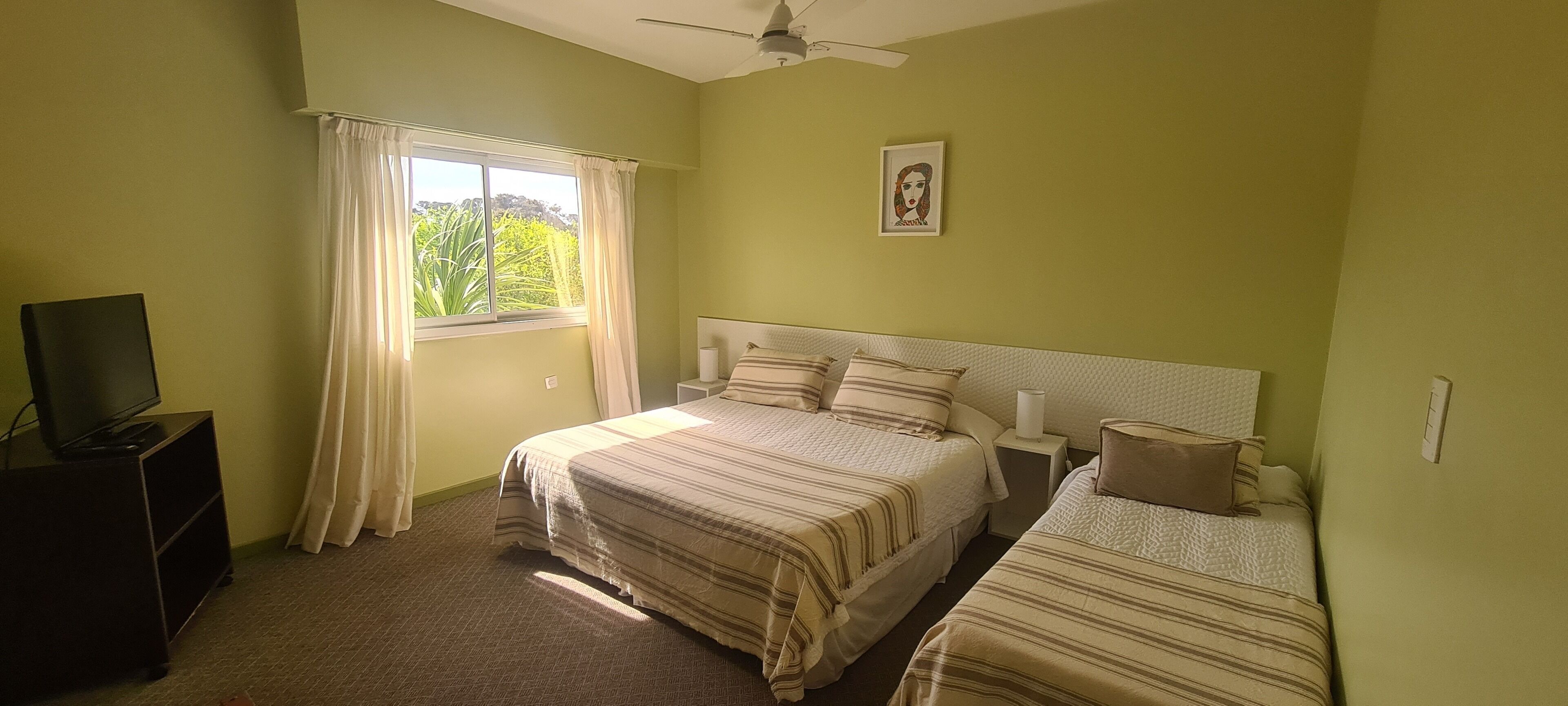 Ein Schlafzimmer mit zwei Betten, einem Deckenventilator, einem Fenster mit Vorhängen, einem gerahmten Bild an der Wand und einem Fernseher auf einem Schrank.