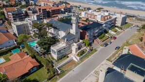 An aerial view of a coastal residential area with a mix of modern and traditional architecture, including a prominent building with a tower, a beach in the background, and a clear road. - Hotel Arte del Rey (Pinamar)