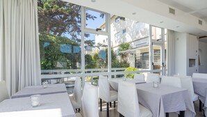 A restaurant with white tables and chairs, large windows, and a view of trees and buildings. - Hotel Arte del Rey (Pinamar)