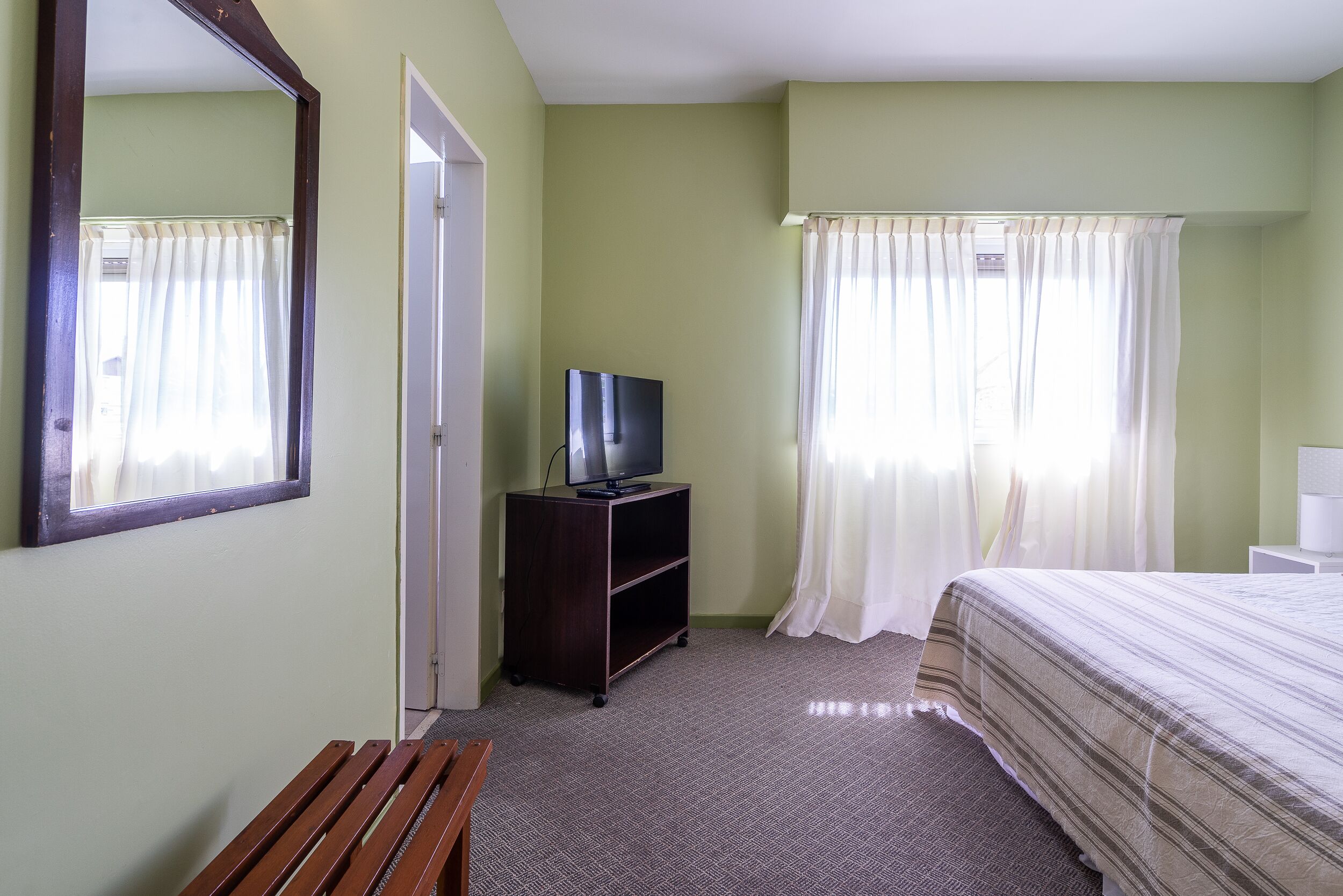 A hotel room with a bed, a TV on a stand, a bench, and a window with curtains.