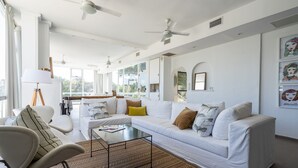 A modern living room with a white sofa, a coffee table, and a chair. There are ceiling fans, large windows, and artwork on the walls. - Hotel Arte del Rey (Pinamar)