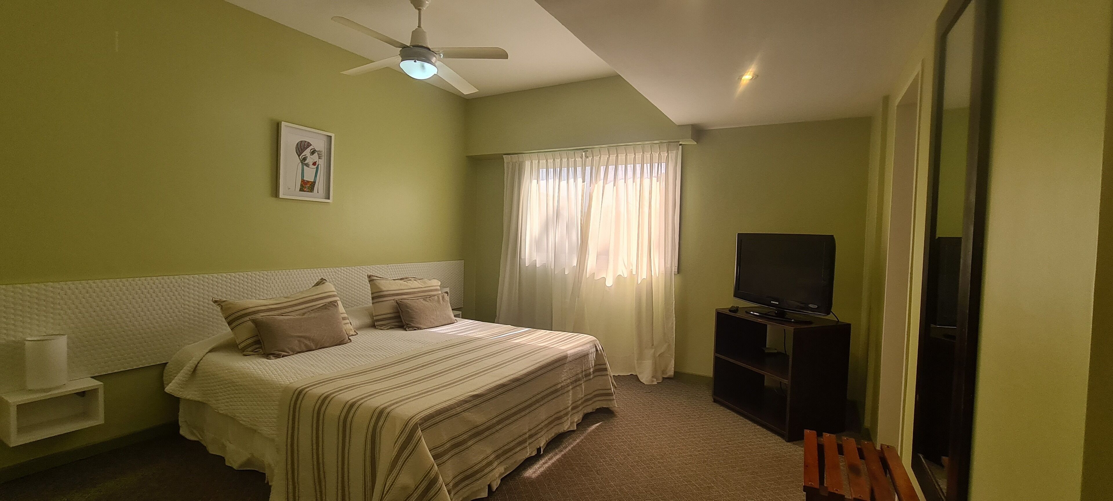 A bedroom with a bed, a ceiling fan, a TV, and a window with curtains.