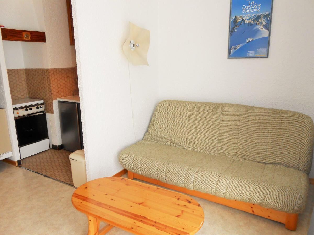 A compact living space with a sofa and a wooden table, a kitchenette with a stove and refrigerator, and a wall-mounted poster.