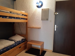 Um quarto com uma cama beliche, uma pequena mesa de madeira, uma luminária na parede e uma porta.