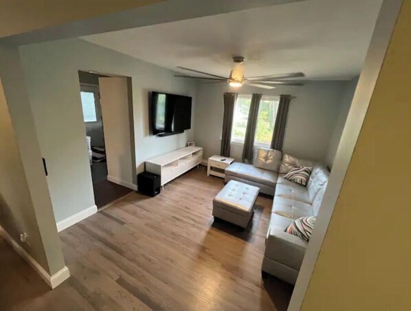 A spacious living room with a ceiling fan, a large flat-screen TV mounted on the wall, a white entertainment unit, a beige sofa, and a wooden floor. - fully Modern 3 bedroom 65"smart tvs cable bluetooth speakers etc (Islip)
