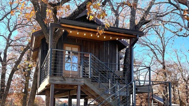 Una cabaña de madera con una escalera que sube a una terraza, rodeada de árboles con hojas otoñales.