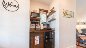 Cocina pequeña con microondas, cafetera y refrigerador. Hay un cartel de 'Welcome' en la pared y un cuadro de un pájaro.