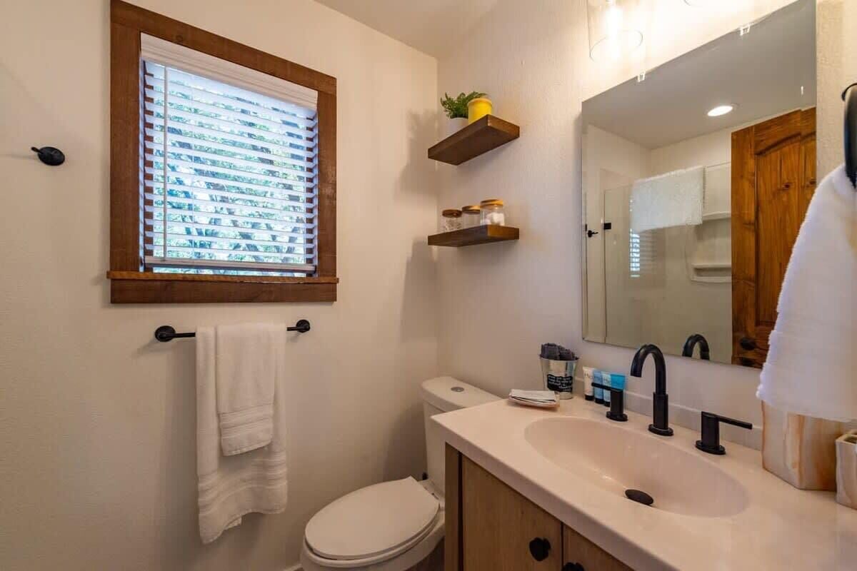 A bathroom with a toilet, sink, and window with blinds. There are wooden shelves above the sink and a towel hanging on a rack.