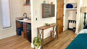A compact living space with a kitchenette, a wooden door, a bed, and a small table with a plant. - Painted Bunting Cabin (15 MIN to Magnolia/Baylor) (Elm Mott)
