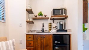 Cocina compacta con armarios de madera, un microondas y un refrigerador. Hay una encimera pequeña con un lavamanos y una cafetera. Un sofá color beige con almohadones a rayas está en primer plano, y una alfombra estampada cubre el suelo.