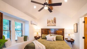 Un dormitorio con cama, ventilador de techo, ventana con persianas, mesita de noche con lámpara y aire acondicionado de pared.