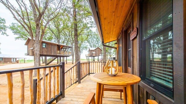A wooden deck with a table and chairs, surrounded by trees and wooden cabins. - The Nuthatch Cabin (15 MIN to Magnolia & Baylor) (Elm Mott)