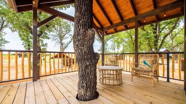 A wooden deck with a pergola, a large tree trunk, wicker chairs, and a view of a fenced area with trees. - The Sparrow Treehouse 15 MIN to Magnolia & Baylor (Elm Mott)