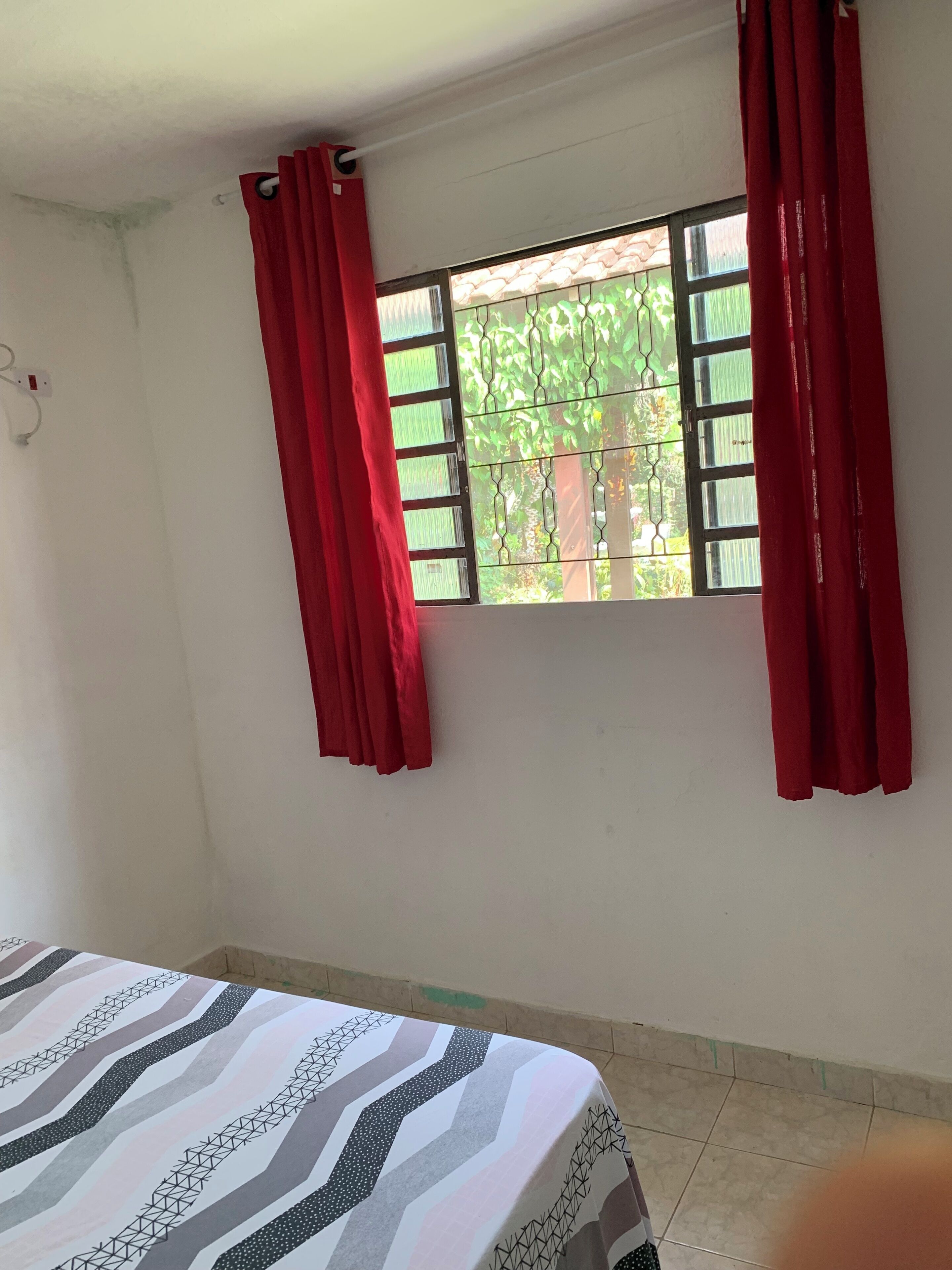 A room with a bed, a window with red curtains, and a view of greenery outside.