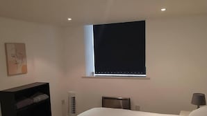 Een slaapkamer met een bed, een nachtkastje met een lamp, een verwarming, een raam met zonwering en een plank met opgevouwen kleding.
