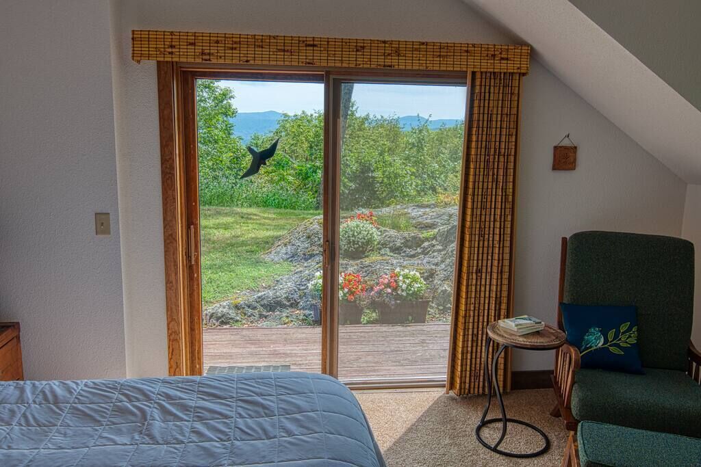 Une chambre avec un lit, une chaise, une petite table et une porte-fenêtre coulissante donnant sur un espace extérieur avec vue sur la verdure et un oiseau.
