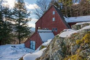 Une cabane en bois rouge, avec un toit en pente, entourée d’un paysage enneigé et d’arbres.