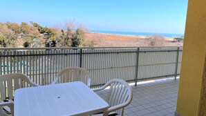 Un balcón con mesa y sillas blancas, baranda de metal y vista al mar y al paisaje.