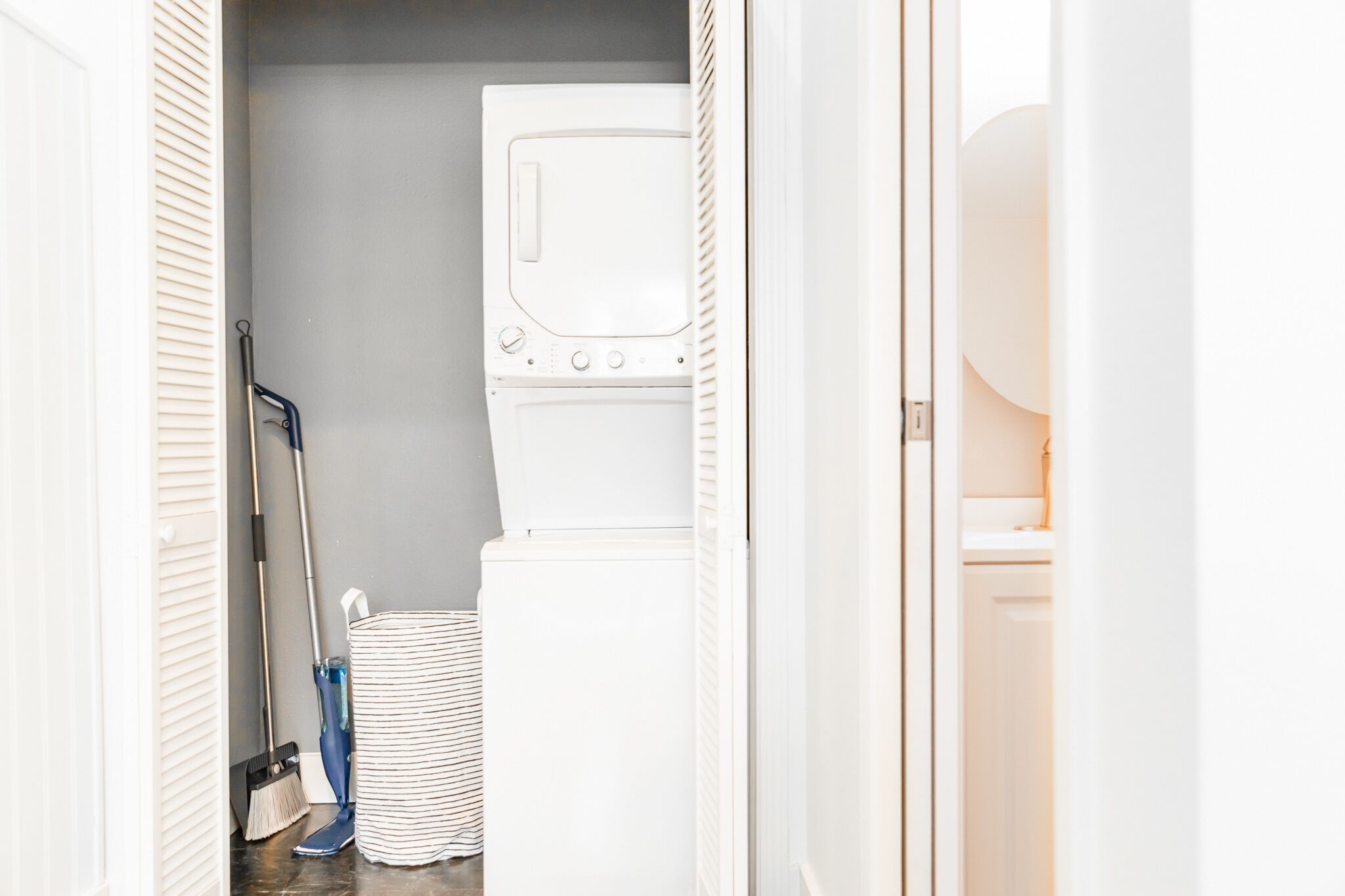 A laundry room with a stacked washer and dryer, a mop, and a striped laundry basket.
