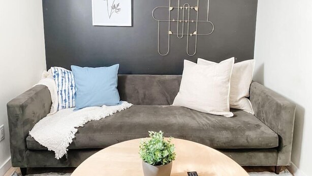 Un canapé gris agrémenté de coussins bleus et blancs, une petite table ronde avec une plante, et une œuvre d’art abstraite accrochée au mur.