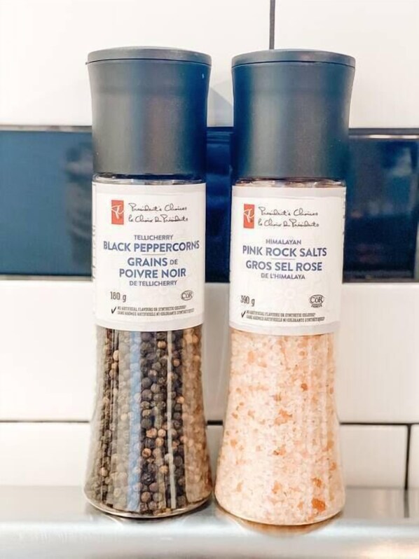 Deux récipients contenant des grains de poivre noir et du sel de roche rose sont posés sur un plan de travail de cuisine.