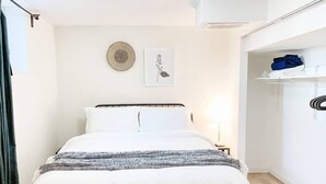 Une chambre à coucher bien rangée, avec un lit, une lampe de chevet, une œuvre d’art accrochée au mur et une étagère où sont pliés des vêtements.