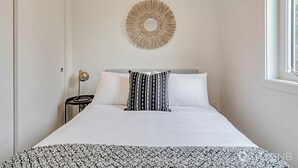 Un lit bien fait, recouvert d’un couvre-lit à motifs, un luminaire rond fixé au mur et une table de chevet avec une lampe.