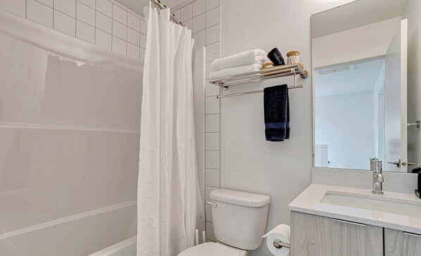 Ein modernes Badezimmer mit Toilette, einer Dusche mit weißem Vorhang, einem Handtuchhalter mit gefalteten Handtüchern und einem Spiegel über dem Waschbecken.