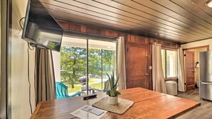 A spacious room with a wooden dining table, a TV mounted on the wall, and a view of the outdoors.