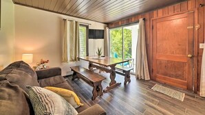 A cozy living room with a wooden ceiling, a large window with curtains, a wooden dining table, and a sofa with cushions.
