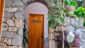 A stone archway leading to a wooden door with a sign, surrounded by potted plants and a tiled floor.