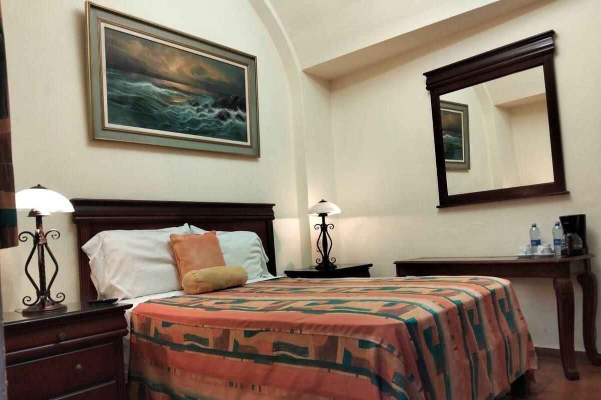 Ein Hotelzimmer mit einem Bett, Nachttischen, Lampen, einem Schreibtisch, einem Spiegel und einem Bild an der Wand.