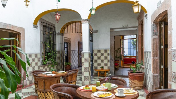 A courtyard with round wooden tables and chairs, surrounded by potted plants and a tiled floor. - Hotel Meson de Isabel (Querétaro)