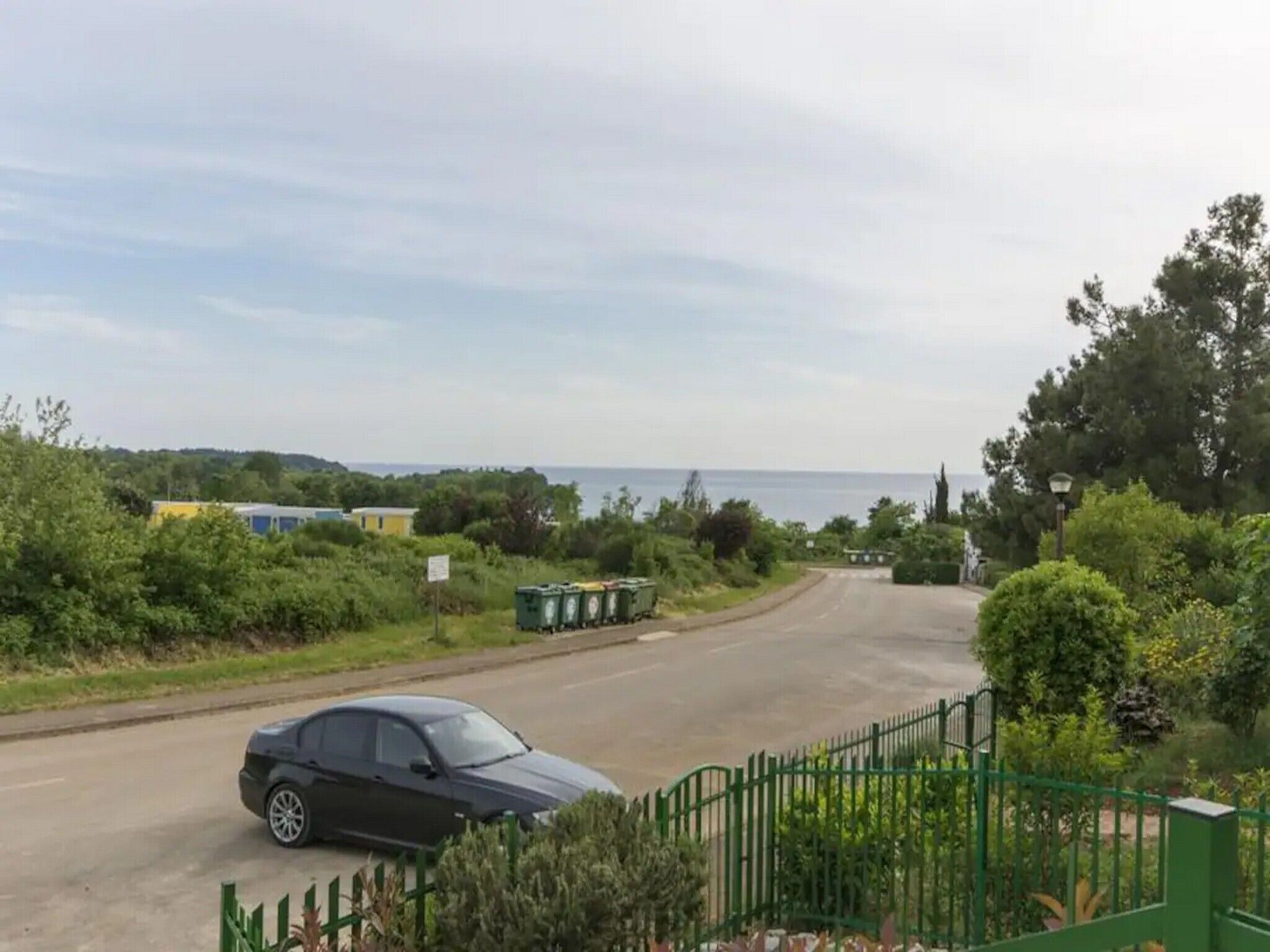 A road with a parked car, green fencing, and a body of water in the background.