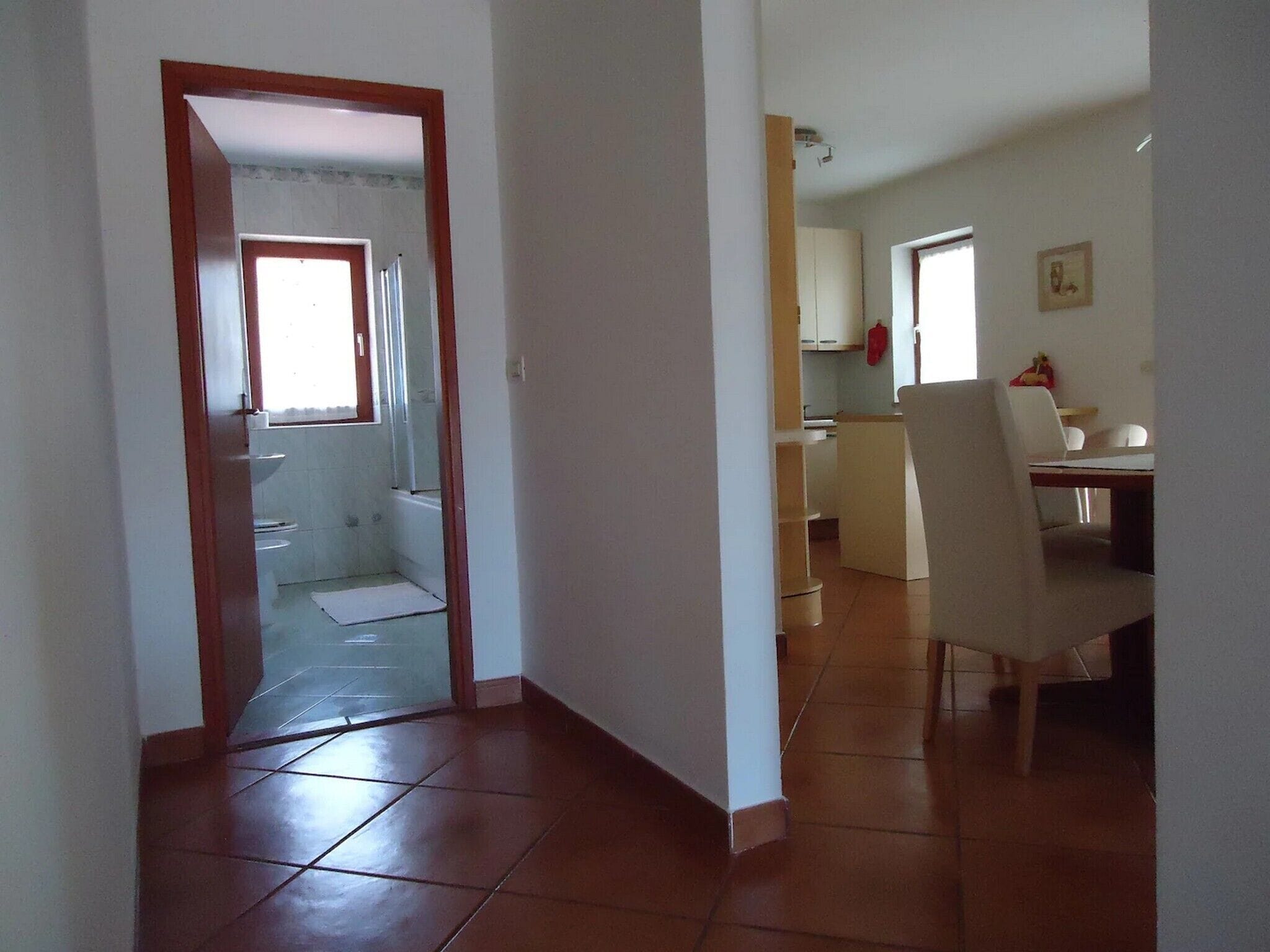 Un pasillo que conduce a un baño con ducha e inodoro, un comedor con mesa y sillas, y una cocina con armarios y un objeto rojo en la pared.
